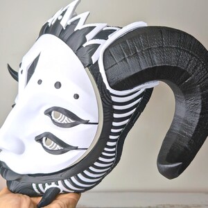 May include: A white and black mask with large, curved horns. The mask features a stylized face with eye details and a decorative crown. The horns are black, and the face is white with black accents. The mask is held in a hand.