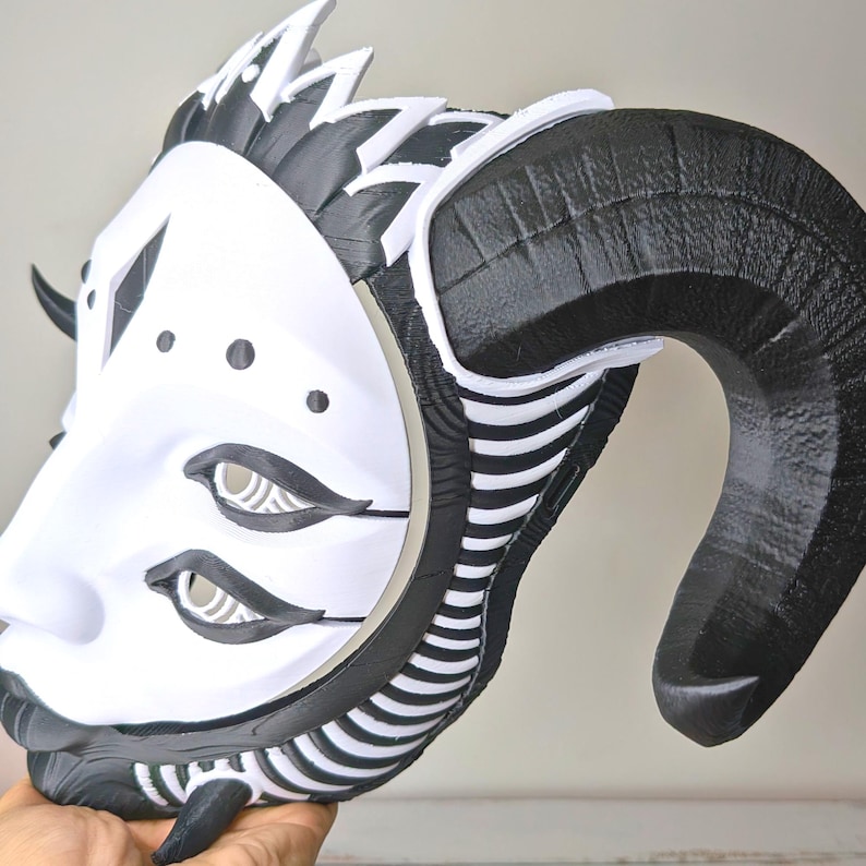 May include: A white and black mask with large, curved horns. The mask features a stylized face with eye details and a decorative crown. The horns are black, and the face is white with black accents. The mask is held in a hand.