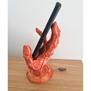 May include: A 3D printed phone stand in the shape of a robotic hand holding a black smartphone. The hand is a bright orange color and has a circular base.