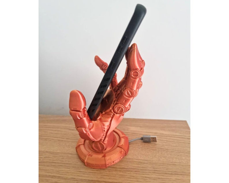 May include: A 3D printed phone stand in the shape of a robotic hand holding a black smartphone. The hand is a bright orange color and has a circular base.