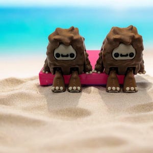 K&ouml;nnte beinhalten: Zwei braune, pelzige Kreaturen mit Comic-Gesichtern sitzen auf einem rosa Strandtuch an einem Sandstrand.