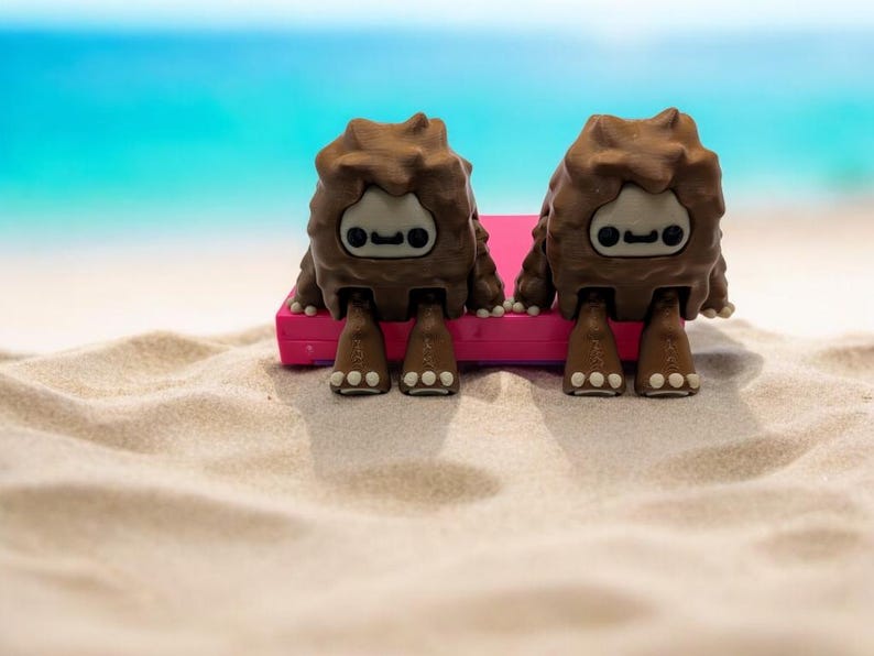 K&ouml;nnte beinhalten: Zwei braune, pelzige Kreaturen mit Comic-Gesichtern sitzen auf einem rosa Strandtuch an einem Sandstrand.