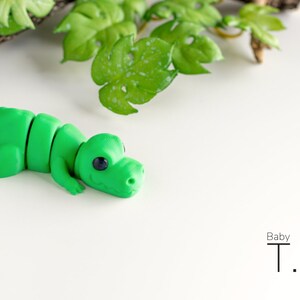 K&ouml;nnte beinhalten: Ein gr&uuml;nes Plastik-Dinosaurierspielzeug, ein Baby T. Rex, liegt auf der Seite auf einer wei&szlig;en Oberfl&auml;che. Der Dinosaurier hat gro&szlig;e Augen und einen kleinen Mund.