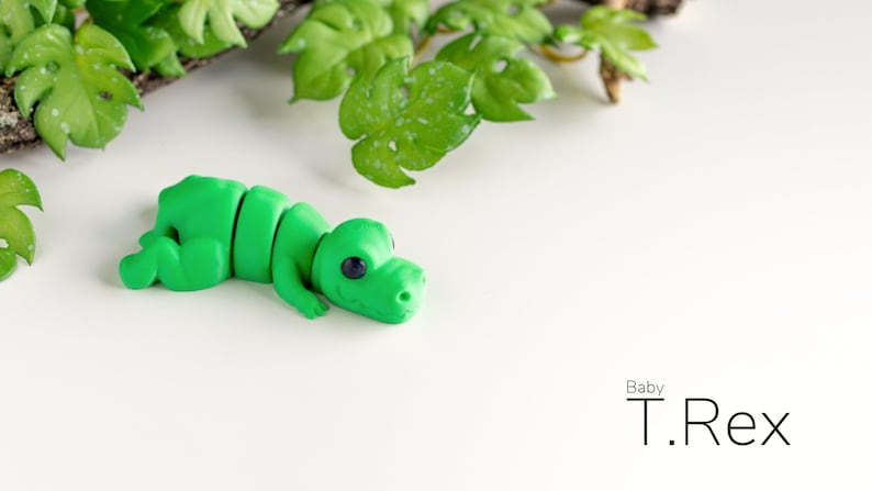 K&ouml;nnte beinhalten: Ein gr&uuml;nes Plastik-Dinosaurierspielzeug, ein Baby T. Rex, liegt auf der Seite auf einer wei&szlig;en Oberfl&auml;che. Der Dinosaurier hat gro&szlig;e Augen und einen kleinen Mund.