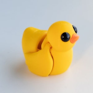 May include: A yellow rubber duck toy with black eyes and an orange beak. The duck is made of multiple pieces that can be moved and rotated.