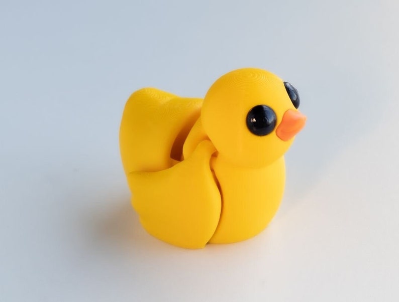 May include: A yellow rubber duck toy with black eyes and an orange beak. The duck is made of multiple pieces that can be moved and rotated.