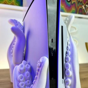 May include: A white 3D printed octopus stand for a video game console. The console is white and black and has a purple glow. The stand is shaped like an octopus with tentacles wrapping around the console.
