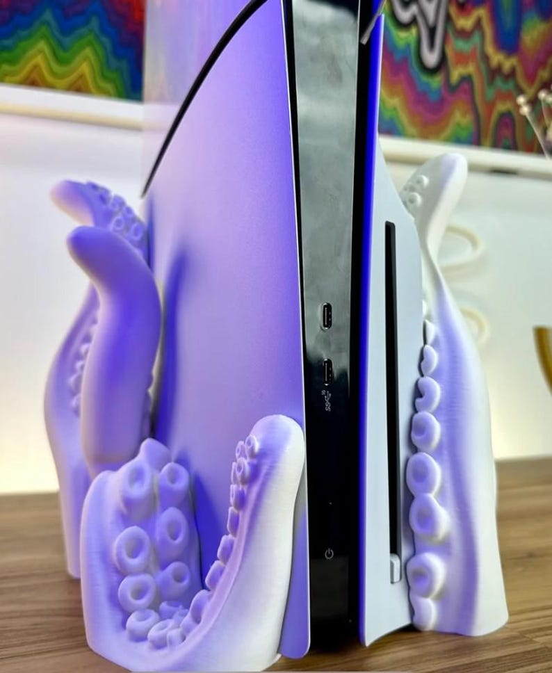 May include: A white 3D printed octopus stand for a video game console. The console is white and black and has a purple glow. The stand is shaped like an octopus with tentacles wrapping around the console.
