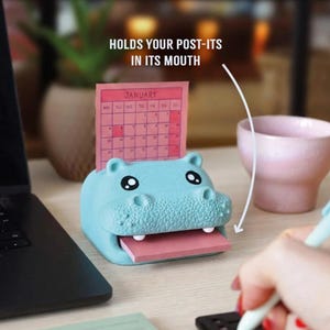 May include: A light blue hippo-shaped desk organizer holding a pink sticky note pad. The hippo has black eyes and a textured snout. A pink calendar is displayed behind the hippo. Text on the image reads "HOLDS YOUR POST-ITS IN ITS MOUTH".