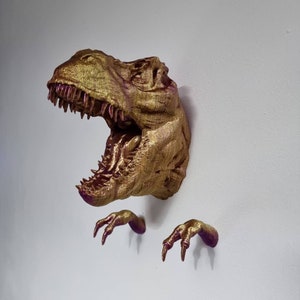 May include: A gold-colored dinosaur head wall mount with a textured finish. The dinosaur has an open mouth with sharp teeth and two separate claws on either side of the head.