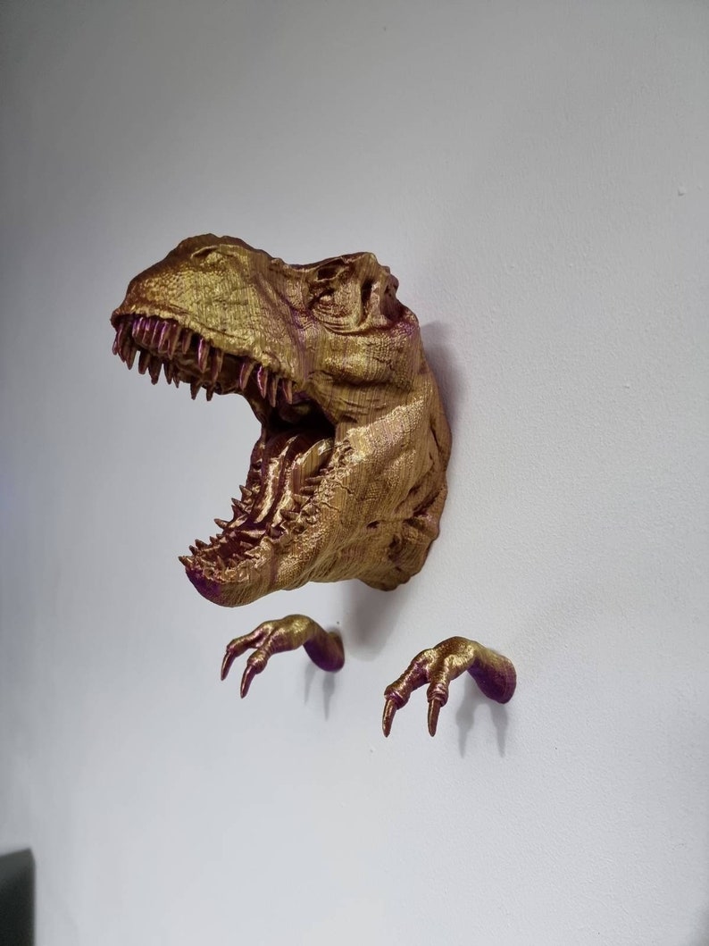 May include: A gold-colored dinosaur head wall mount with a textured finish. The dinosaur has an open mouth with sharp teeth and two separate claws on either side of the head.