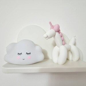 May include: A white shelf with a white cloud-shaped nightlight with a smiling face and closed eyes and a white balloon animal unicorn with pink accents.