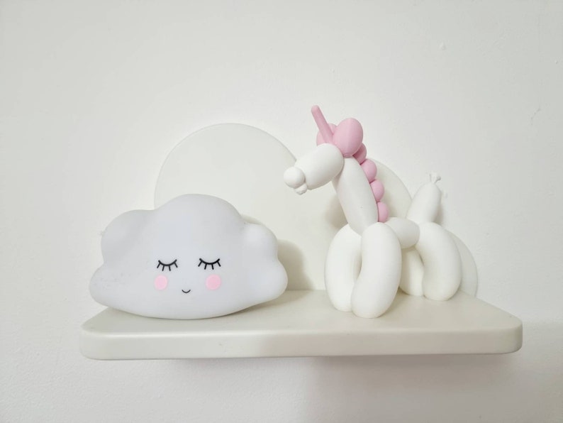 May include: A white shelf with a white cloud-shaped nightlight with a smiling face and closed eyes and a white balloon animal unicorn with pink accents.