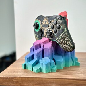 May include: A black and green video game controller with gold accents sits on a colorful, multi-colored, 3D printed stand. The controller has a geometric pattern and the stand is shaped like a mountain range.
