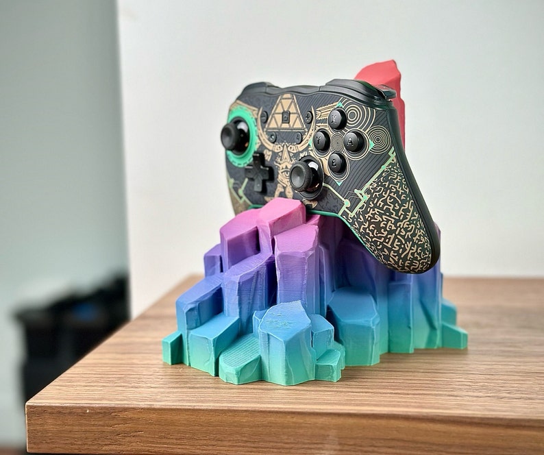 May include: A black and green video game controller with gold accents sits on a colorful, multi-colored, 3D printed stand. The controller has a geometric pattern and the stand is shaped like a mountain range.