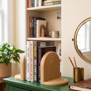 Op de afbeelding: Beige boogvormige boekensteunen met een gelaagd ontwerp houden verschillende boeken vast. De boeken hebben titels als "How I Built This" en "Limitless". Een potplant, spiegel en potloodhouder zijn ook zichtbaar.