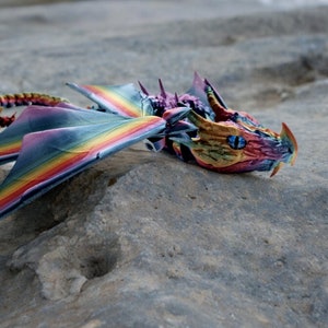 May include: A rainbow-coloured dragon figurine with blue eyes, lying on a grey rock surface.