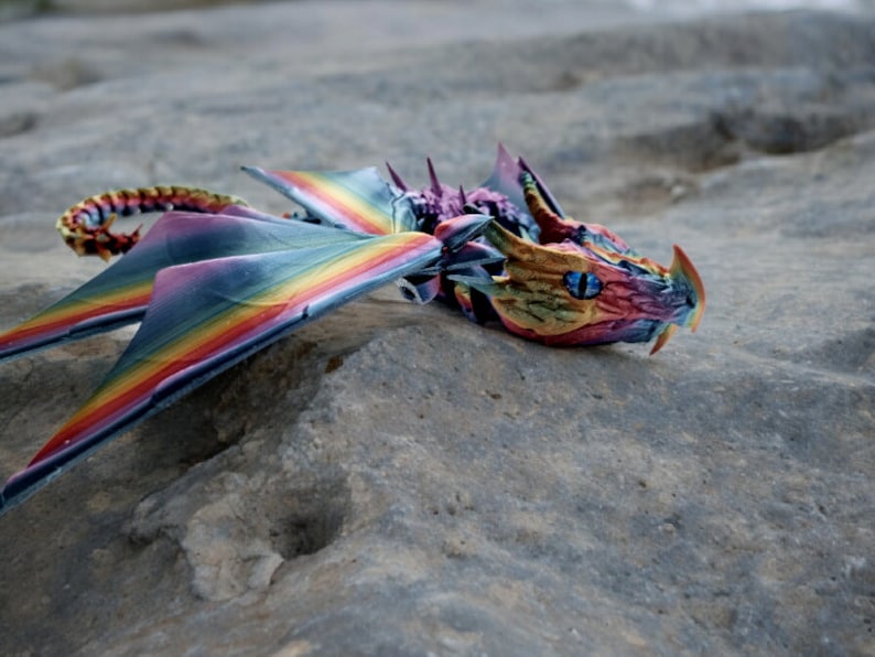 May include: A rainbow-coloured dragon figurine with blue eyes, lying on a grey rock surface.