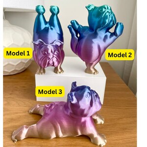 K&ouml;nnte beinhalten: Drei Bulldoggen-Figuren in verschiedenen Posen, jede mit einem Farbverlauf aus Blau, Lila und Gold. Modell 1 steht auf dem Kopf, Modell 2 hat die Arme erhoben und Modell 3 liegt. Die Modelle sind mit gelben Rechtecken beschriftet.