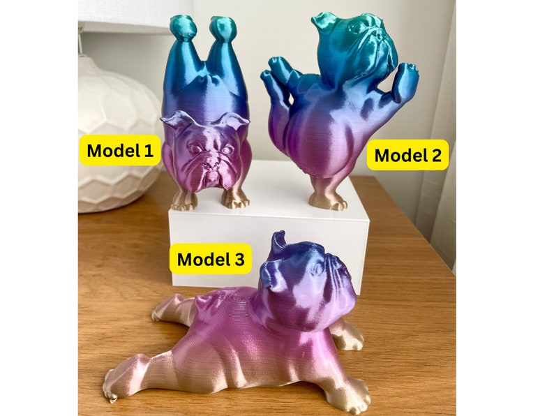 K&ouml;nnte beinhalten: Drei Bulldoggen-Figuren in verschiedenen Posen, jede mit einem Farbverlauf aus Blau, Lila und Gold. Modell 1 steht auf dem Kopf, Modell 2 hat die Arme erhoben und Modell 3 liegt. Die Modelle sind mit gelben Rechtecken beschriftet.
