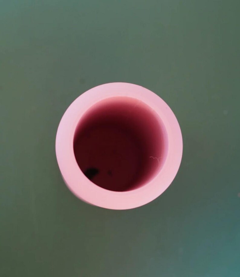 May include: A top-down view of a light pink, circular object with a hollow center. The inside of the object is a darker shade of pink. The object is set against a muted green background.