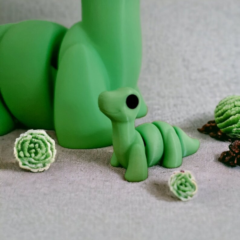 Pu&ograve; includere: Un dinosauro di cartone animato verde segmentato con un solo occhio e una piccola bocca. Il dinosauro &egrave; seduto su una superficie grigio chiaro.