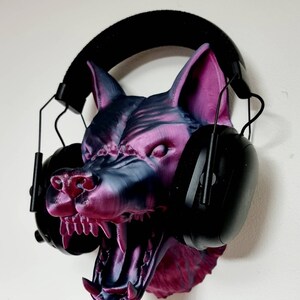 May include: A 3D-printed wolf head in shades of purple and gray, with an open mouth and visible teeth. Black earmuffs are placed over the head. The wolf head is mounted on a white wall.