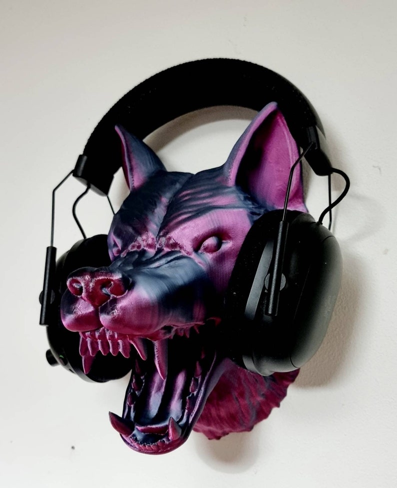 May include: A 3D-printed wolf head in shades of purple and gray, with an open mouth and visible teeth. Black earmuffs are placed over the head. The wolf head is mounted on a white wall.