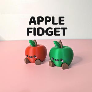 May include: Two apple-shaped fidget toys, one red and one green, with smiling faces and brown feet. The toys have brown stems and green leaves. The text "APPLE FIDGET" is above the toys.