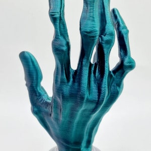 May include: A teal blue 3D printed skeletal hand sculpture with a silver base. The hand is posed with the fingers extended upwards.