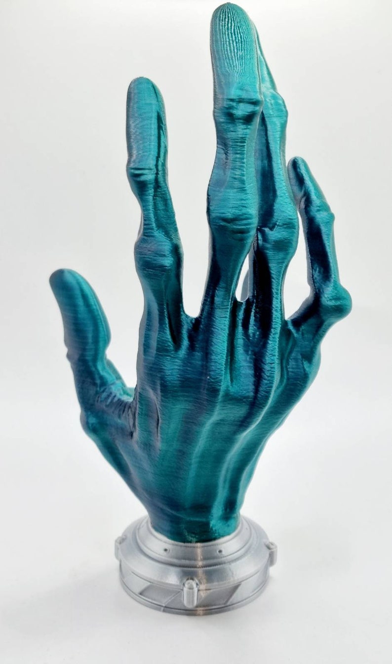 May include: A teal blue 3D printed skeletal hand sculpture with a silver base. The hand is posed with the fingers extended upwards.