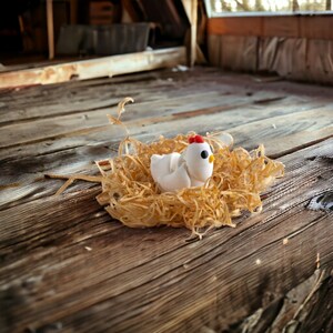 May include: A white ceramic chicken figurine sitting in a nest of shredded paper. The chicken has a red comb and black eyes.