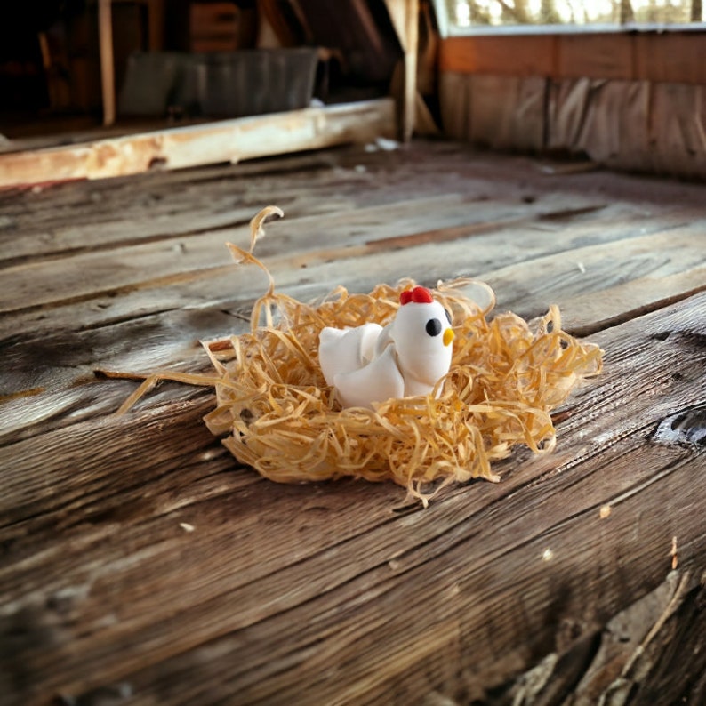 May include: A white ceramic chicken figurine sitting in a nest of shredded paper. The chicken has a red comb and black eyes.
