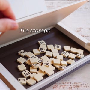 Puede incluir: Una caja de almacenamiento de baldosas blanca y marr&oacute;n con tapa abatible, llena de baldosas de letras beige. El texto "Tile storage" es visible en la tapa. Las baldosas est&aacute;n esparcidas dentro de la caja.
