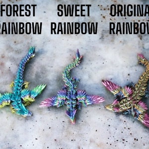 May include: Three origami dragons made of iridescent paper. The dragons are in different color schemes: forest rainbow, sweet rainbow, and original rainbow. The dragons are all facing the same direction with their wings spread out.