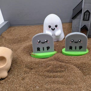 Pu&ograve; includere: Una scena di cimitero in miniatura con due lapidi grigie che recitano "RIP" e hanno facce carine. C'&egrave; una statuetta di fantasma bianco e una statuetta di teschio marrone nella sabbia.