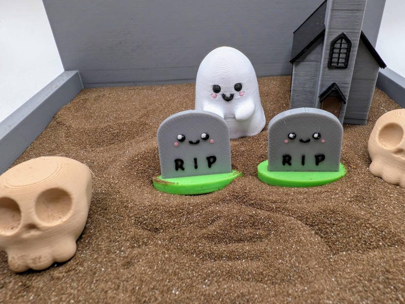 Pu&ograve; includere: Una scena di cimitero in miniatura con due lapidi grigie che recitano "RIP" e hanno facce carine. C'&egrave; una statuetta di fantasma bianco e una statuetta di teschio marrone nella sabbia.