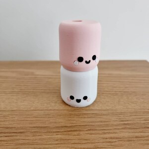 Op de afbeelding: Twee gestapelde keramische figuurtjes in de vorm van een marshmallow met geschilderde gezichten. Het bovenste figuurtje is roze en het onderste figuurtje is wit.