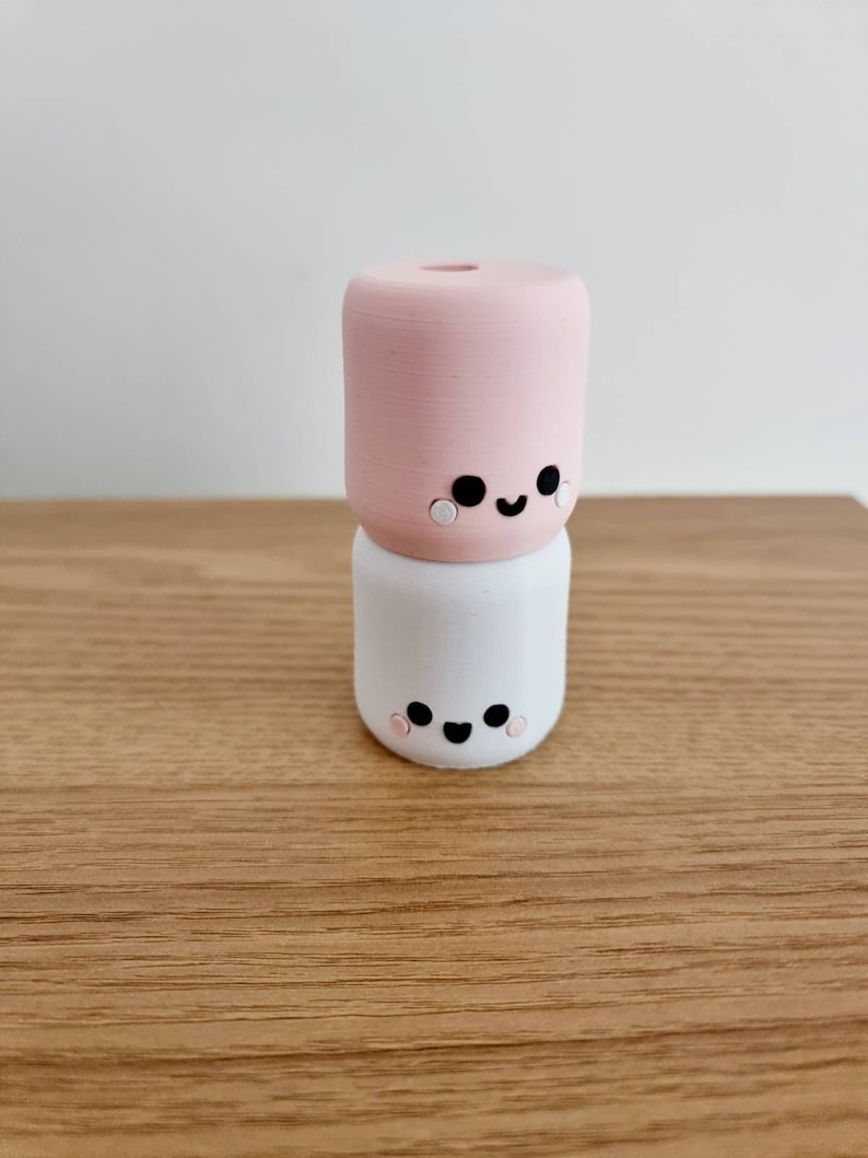 Op de afbeelding: Twee gestapelde keramische figuurtjes in de vorm van een marshmallow met geschilderde gezichten. Het bovenste figuurtje is roze en het onderste figuurtje is wit.