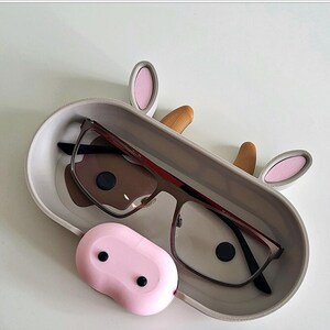 May include: A gray cow-shaped glasses holder with pink ears and a brown nose. The holder has a gray interior and a pair of glasses resting inside. The holder has a pink, rounded, cow udder-shaped compartment on the bottom.