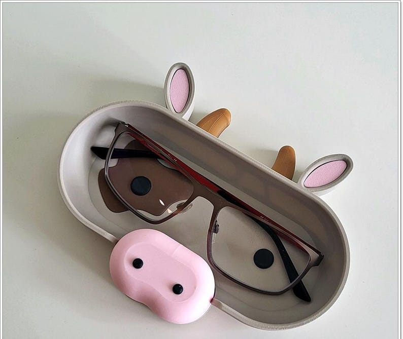 May include: A gray cow-shaped glasses holder with pink ears and a brown nose. The holder has a gray interior and a pair of glasses resting inside. The holder has a pink, rounded, cow udder-shaped compartment on the bottom.