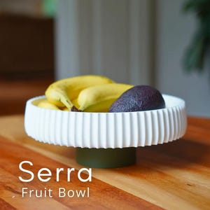 Puede incluir: Cuenco de fruta de cer&aacute;mica blanca con dise&ntilde;o acanalado, con pl&aacute;tanos y un aguacate. El cuenco se apoya sobre una base verde. El texto "Serra Fruit Bowl" se muestra debajo.