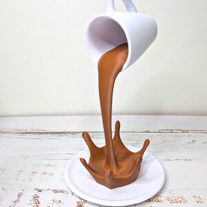 May include: A white ceramic mug appears to be pouring a stream of brown liquid, forming a splash on a white plate. The mug is suspended in mid-air, creating an optical illusion. The background is a light-colored wooden surface.