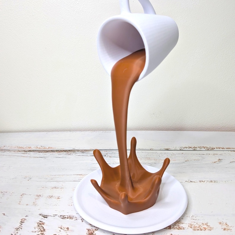May include: A white ceramic mug appears to be pouring a stream of brown liquid, forming a splash on a white plate. The mug is suspended in mid-air, creating an optical illusion. The background is a light-colored wooden surface.