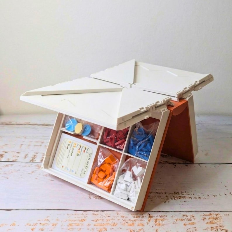 May include: An open board game storage box with a white lid and orange sides. The box is divided into compartments containing game pieces in red, blue, orange, and white. The game pieces are in clear plastic bags. A game card is visible.