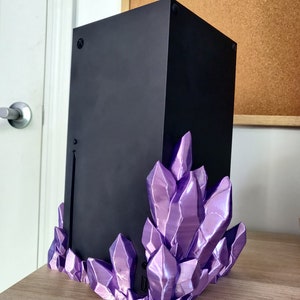 Puede incluir: Una consola de juegos negra con un soporte en forma de cristal morado. La consola est&aacute; sentada sobre una superficie de madera.