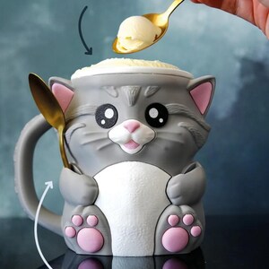 May include: A gray cat-shaped mug with pink accents, holding a golden spoon. The mug has a white belly and pink paw prints. Text on the image reads "FITS A REGULAR PINT" and "AND HOLDS TWO SPOONS". The mug is filled with ice cream.