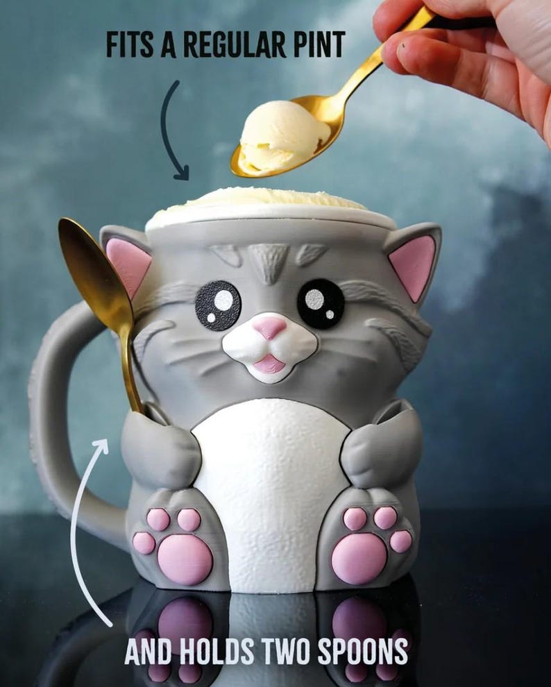 May include: A gray cat-shaped mug with pink accents, holding a golden spoon. The mug has a white belly and pink paw prints. Text on the image reads "FITS A REGULAR PINT" and "AND HOLDS TWO SPOONS". The mug is filled with ice cream.