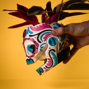 May include: A white skull-shaped planter with a colorful floral design in pink, blue, and black. The planter holds a plant with dark red and green leaves. The planter is held against a yellow background.