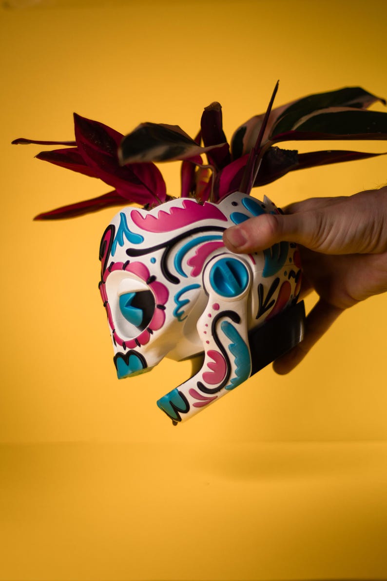 May include: A white skull-shaped planter with a colorful floral design in pink, blue, and black. The planter holds a plant with dark red and green leaves. The planter is held against a yellow background.